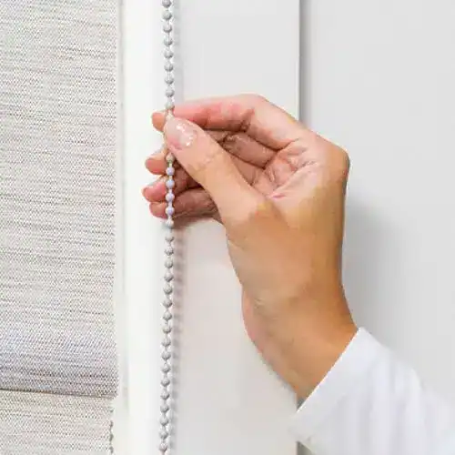 Close-up of a hand adjusting a vertical beaded pull chain for window blinds or curtains. The hand appears delicate and precise, set against a neutral background.