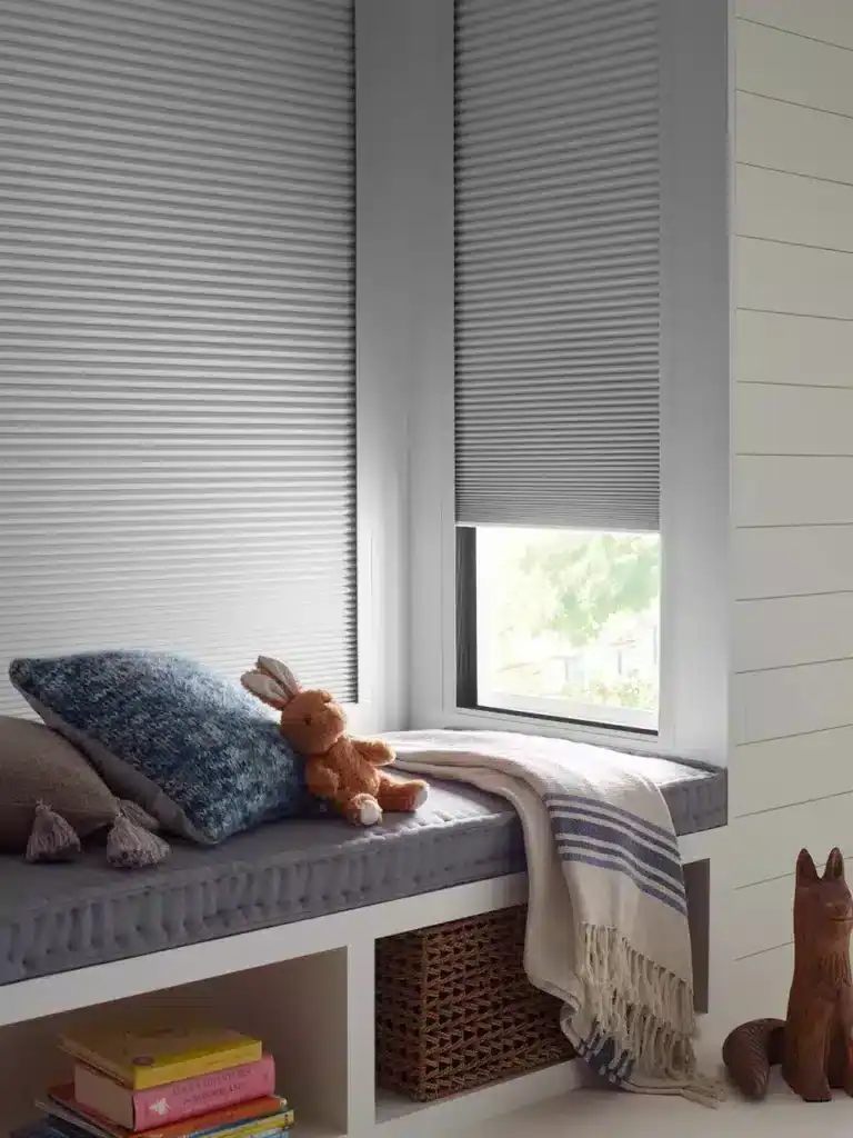 A charming window seat featuring a plush bunny toy, cozy pillows, and a striped blanket, illuminated by natural light through partially closed blinds, with decorative wooden cat and storage beneath.