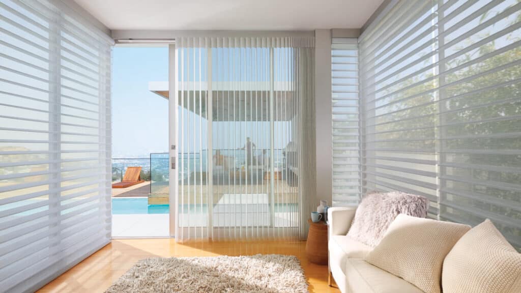 Bright, airy living space featuring expansive windows, vertical blinds, cozy sofa, and a view of the balcony and outdoor pool area.