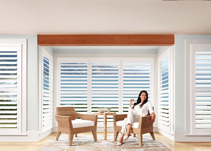 Spacious sunroom with large windows, white shutters, comfortable chairs, and a woman relaxing, surrounded by bright natural sunlight and a scenic view.