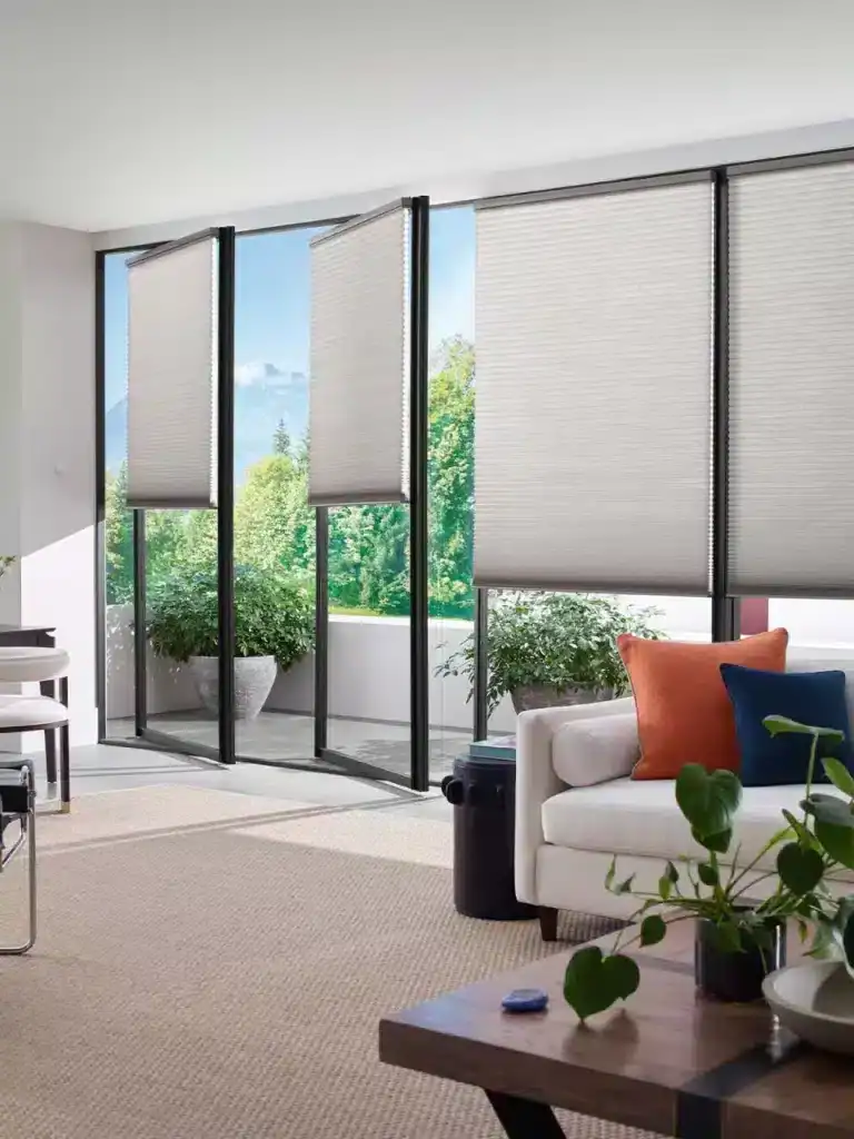 Bright living space featuring large windows with beige blinds and lush green outdoor view.