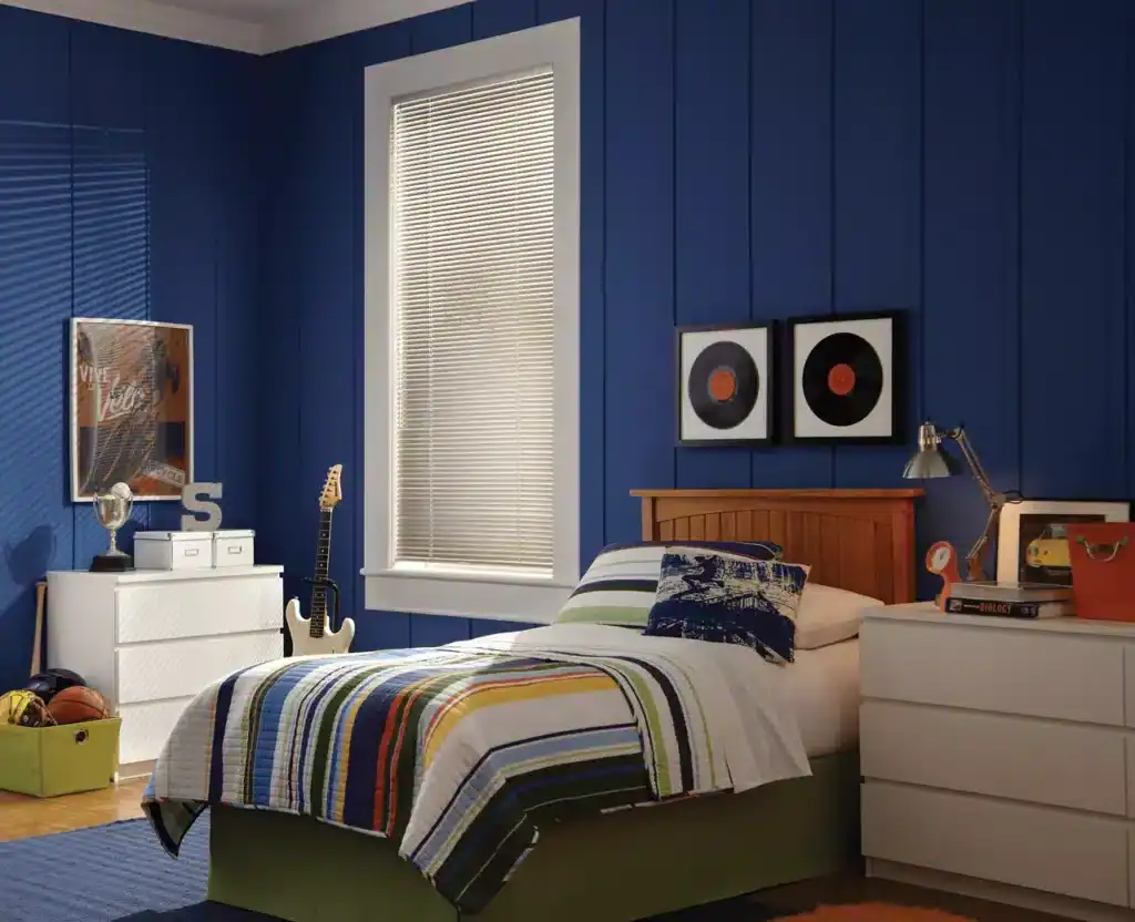 Bright blue bedroom wall with vinyl record art, a white dresser, skateboard, and sports gear, creating a vibrant and lively ambiance. Natural light filters through blinds, illuminating the colorful bedrooms' decor perfect for a creative personality.