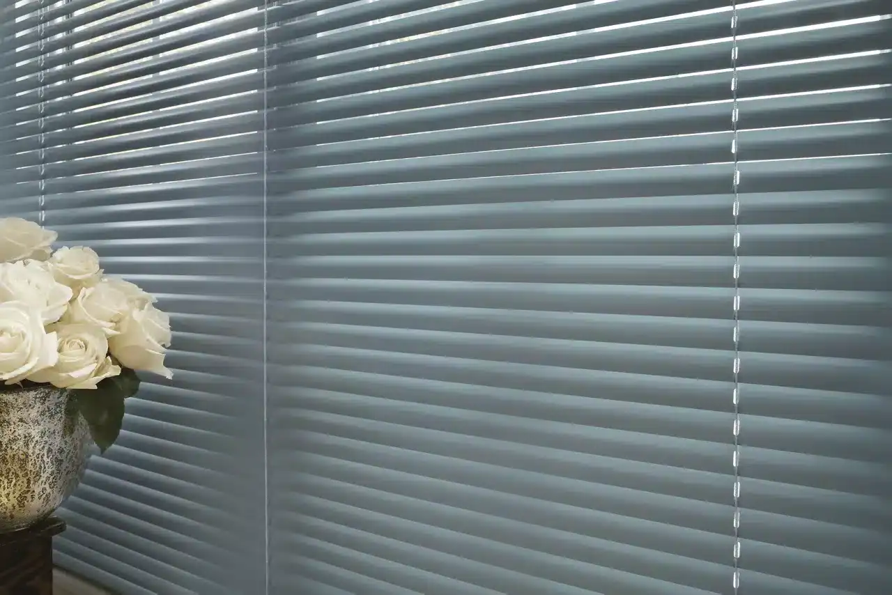 Close-up of beige decorative flower pot filled with white roses in front of window blinds.