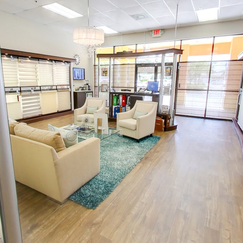 Contemporary office waiting area with beige sofas, white armchairs, glass coffee table, and large windows letting in natural light.