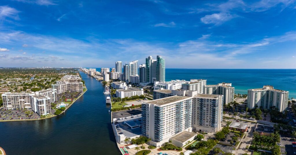 Aerial view of Broward County with high-rise buildings, waterways, and the Atlantic Ocean in the background under a clear blue sky.