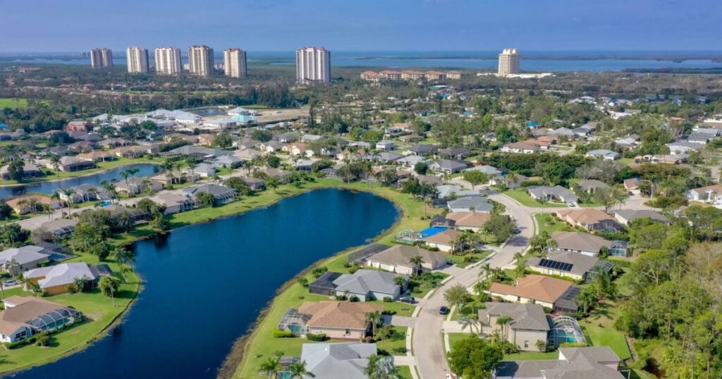 Aerial view of a suburban neighborhood with a lake, surrounded by houses, greenery, and high-rise buildings in the distance, under a blue sky.