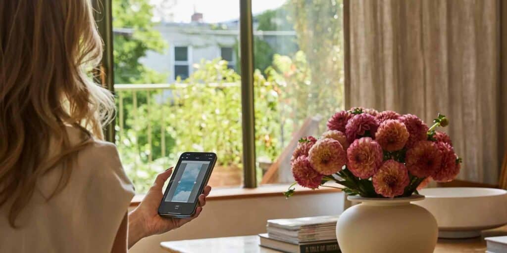 Woman using smartphone to operate motorized drapery in a bright living room with large window and floral arrangement.