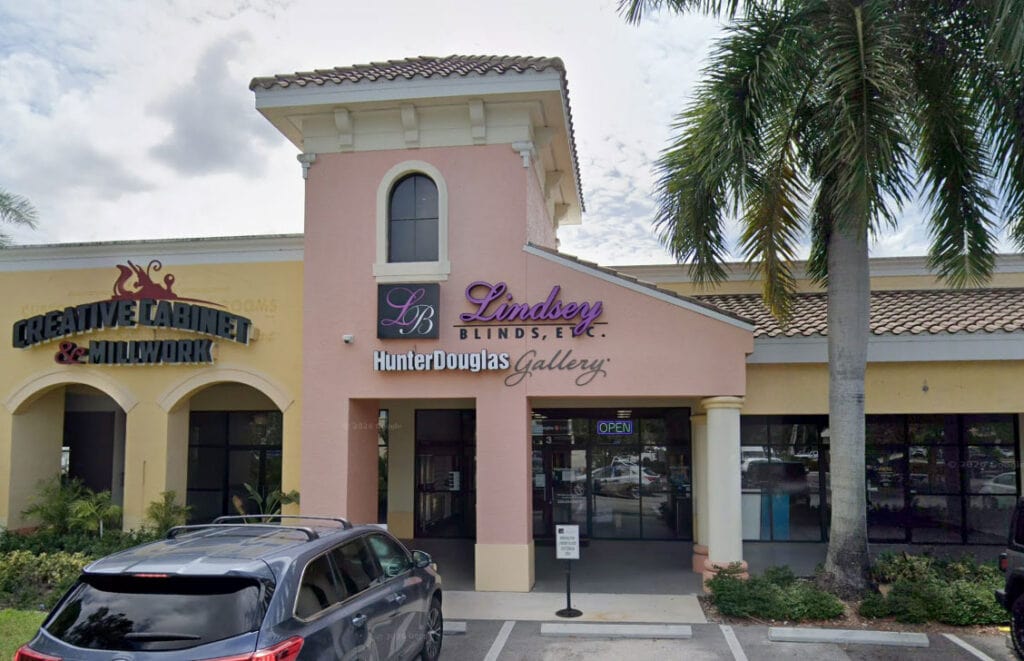 Exterior view of Lindsey Blinds store in Naples with pink facade, palm trees, and parked cars, showcasing custom window treatments and local shopping.