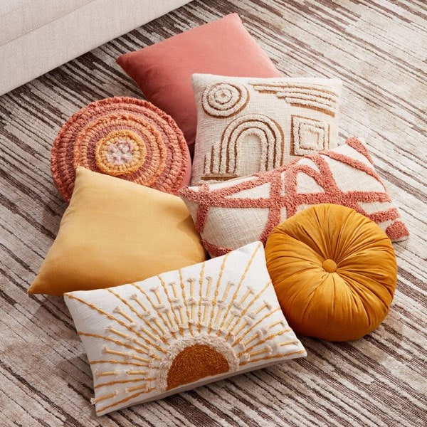 A variety of decorative pillows in pink, yellow, white, and orange with different textures and patterns on a striped rug.