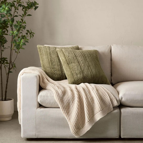 Comfortable beige sofa with two green textured pillows and a cream knitted throw blanket, next to a potted plant, in a cozy living room setting.