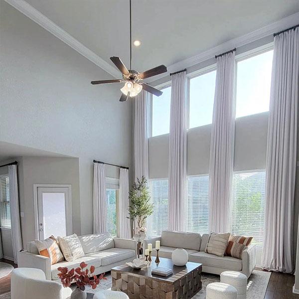 Spacious living room with high ceilings, large windows with white curtains, and modern furniture including white sofas and a wooden coffee table.