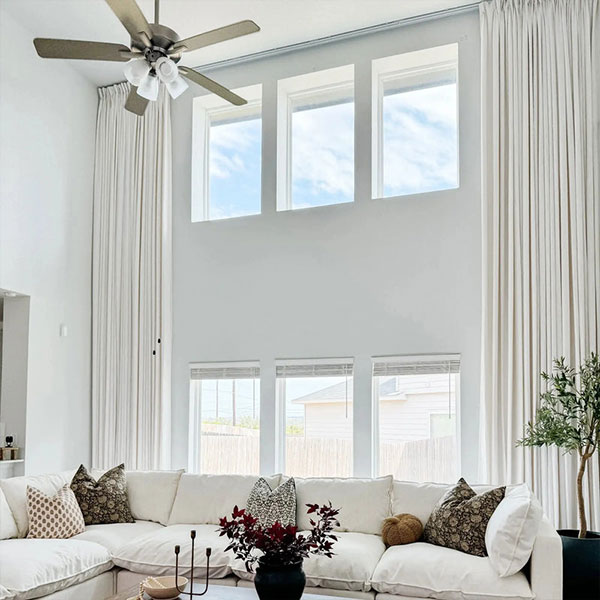 Spacious living room with tall windows, white walls, and a ceiling fan, featuring a large white sectional sofa with decorative pillows and a potted plant.