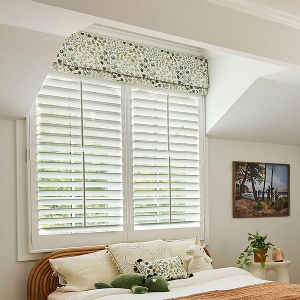Bright room with large white window shutters, floral valance, and cozy bed with plush toys and decorative pillows.