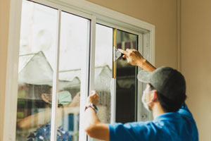 Person cleaning window glass with squeegee, wearing a mask and cap, in a bright room with a view outside.
