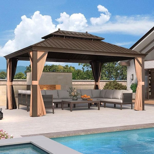 Elegant outdoor gazebo with beige curtains, surrounding a comfortable seating area, overlooking a swimming pool under a blue sky with clouds.